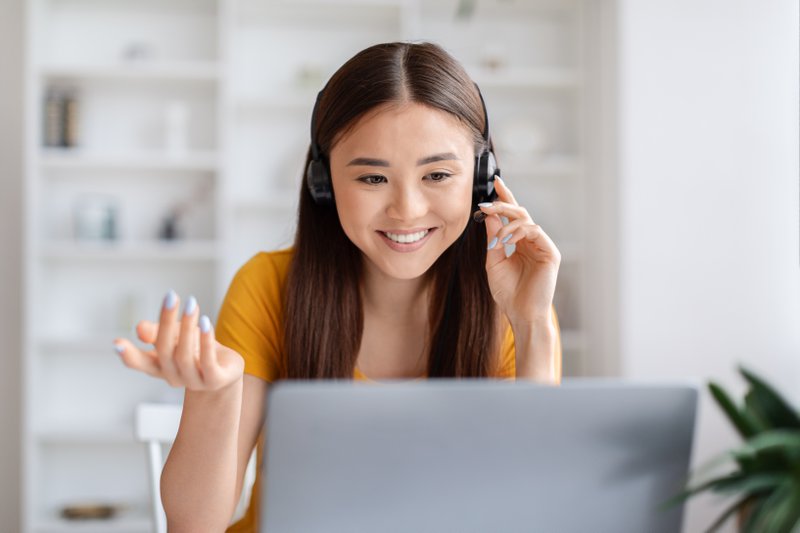Helpful asian customer service representative wearing headset and looking at laptop screen, beautiful korean lady smiling while assisting client over video call, working in home office setting