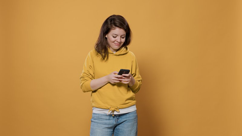 Portrait of casual woman holding smartphone texting having captivating online conversation on social media messaging app in studio. Smiling person in her late 20s using digital touchscreen device.